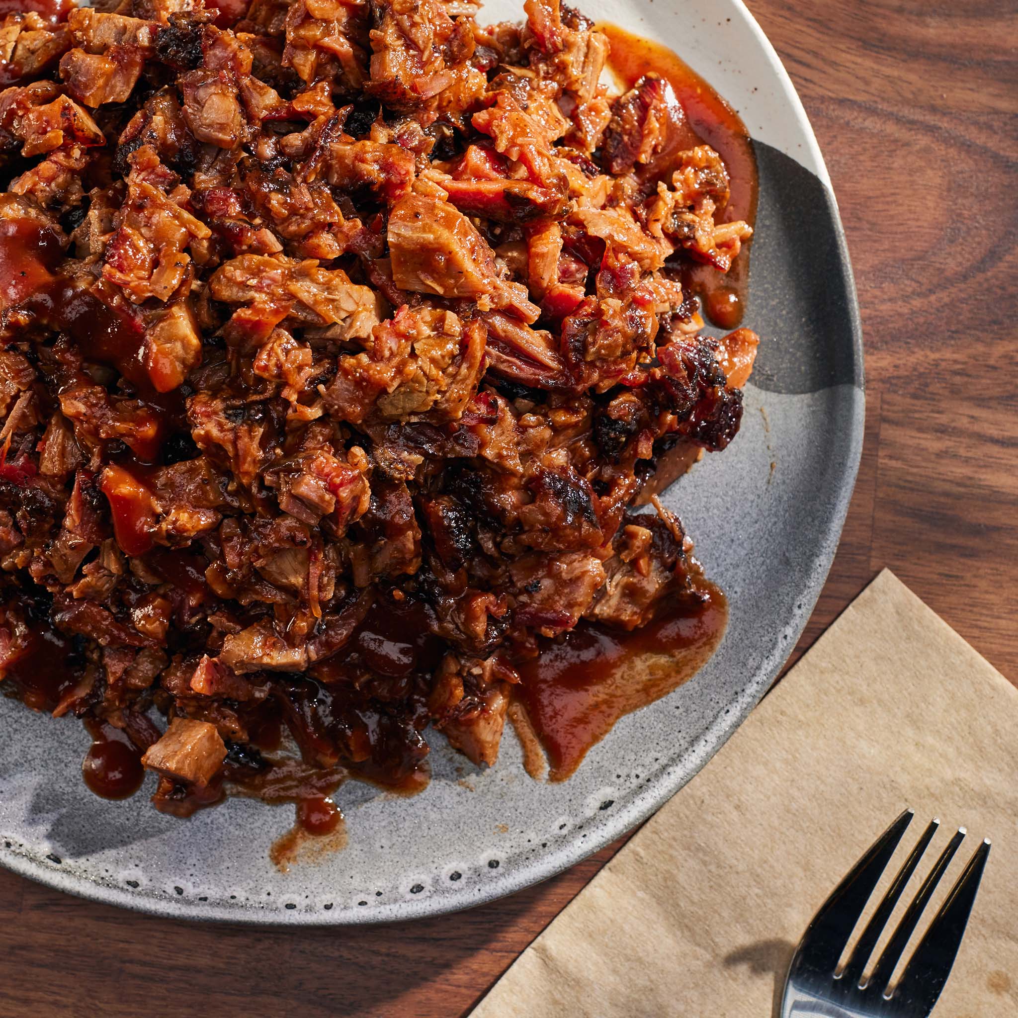 Plated dish of shredded meat with sauce on a wooden table with a fork and napkin.