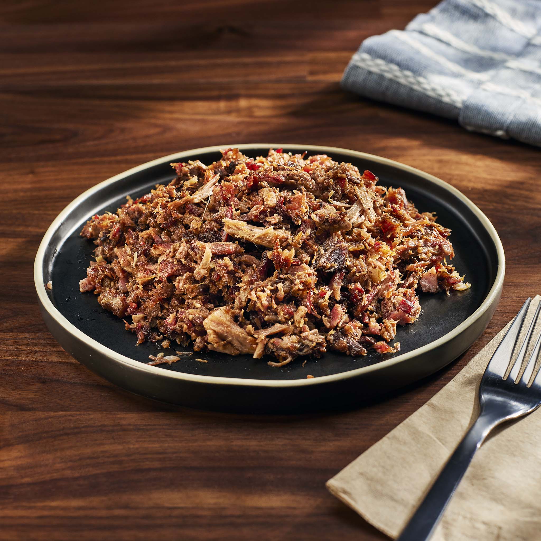 Plated dish of pulled pork on a wooden table with a napkin and fork.