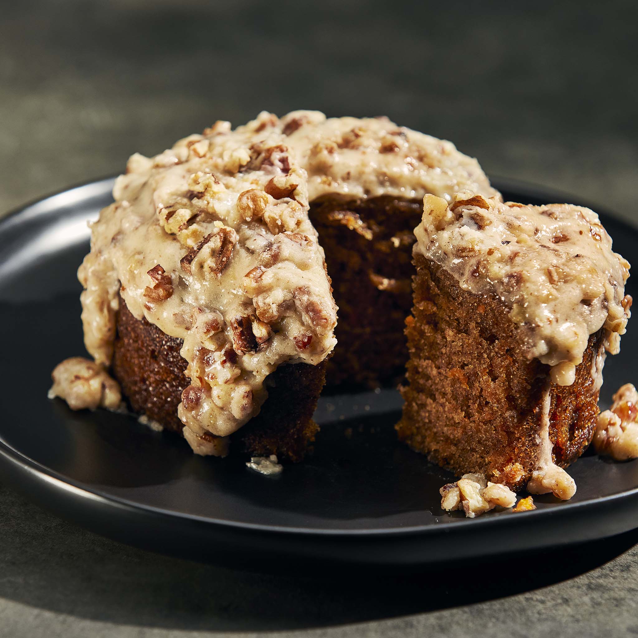 Carrot cake with cream cheese frosting and nuts on a black plate against a dark background