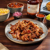 Plated pulled pork with Jack Stack Barbecue sauce on a wooden table with sides and a bottle of sauce.