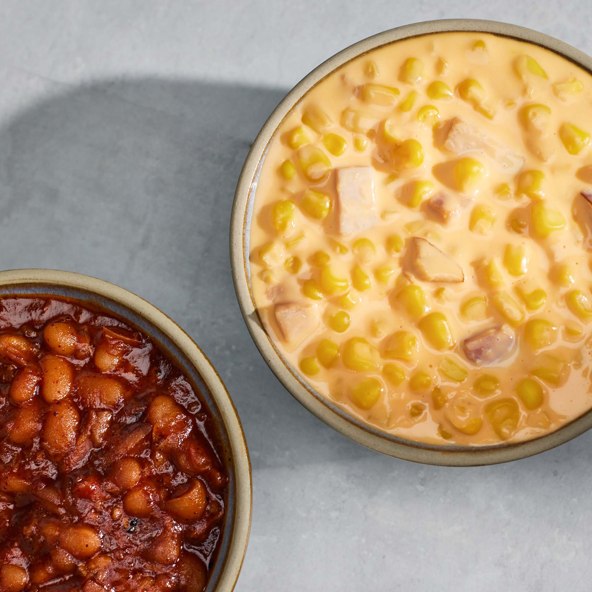 Two bowls of food on a gray surface, one with cheesy corn and the other with beans.