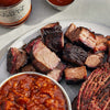 Plated barbecue meats with a side of baked beans and a bottle of barbecue sauce on a light surface.