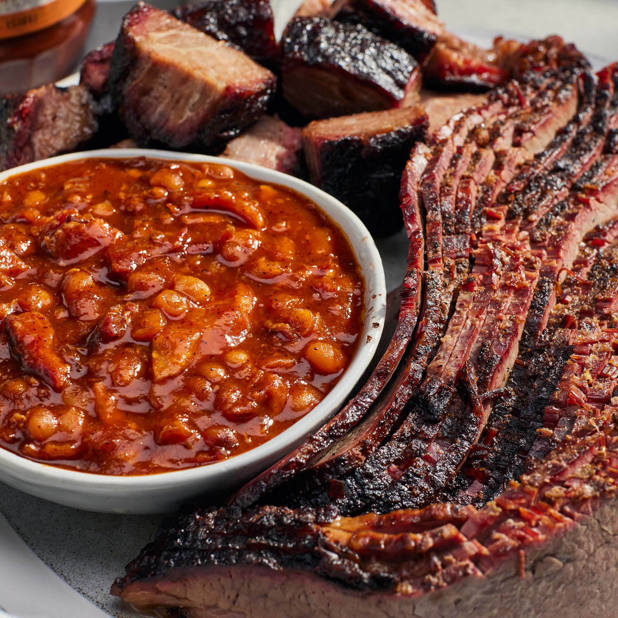 Bowl of baked beans with sliced smoked meat on a wooden cutting board