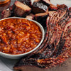 Bowl of baked beans with sliced smoked meat on a wooden cutting board