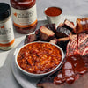 Platter of smoked meats with a bowl of baked beans and two bottles of Fiedler's Jack Stack Barbecue sauce on a gray surface.