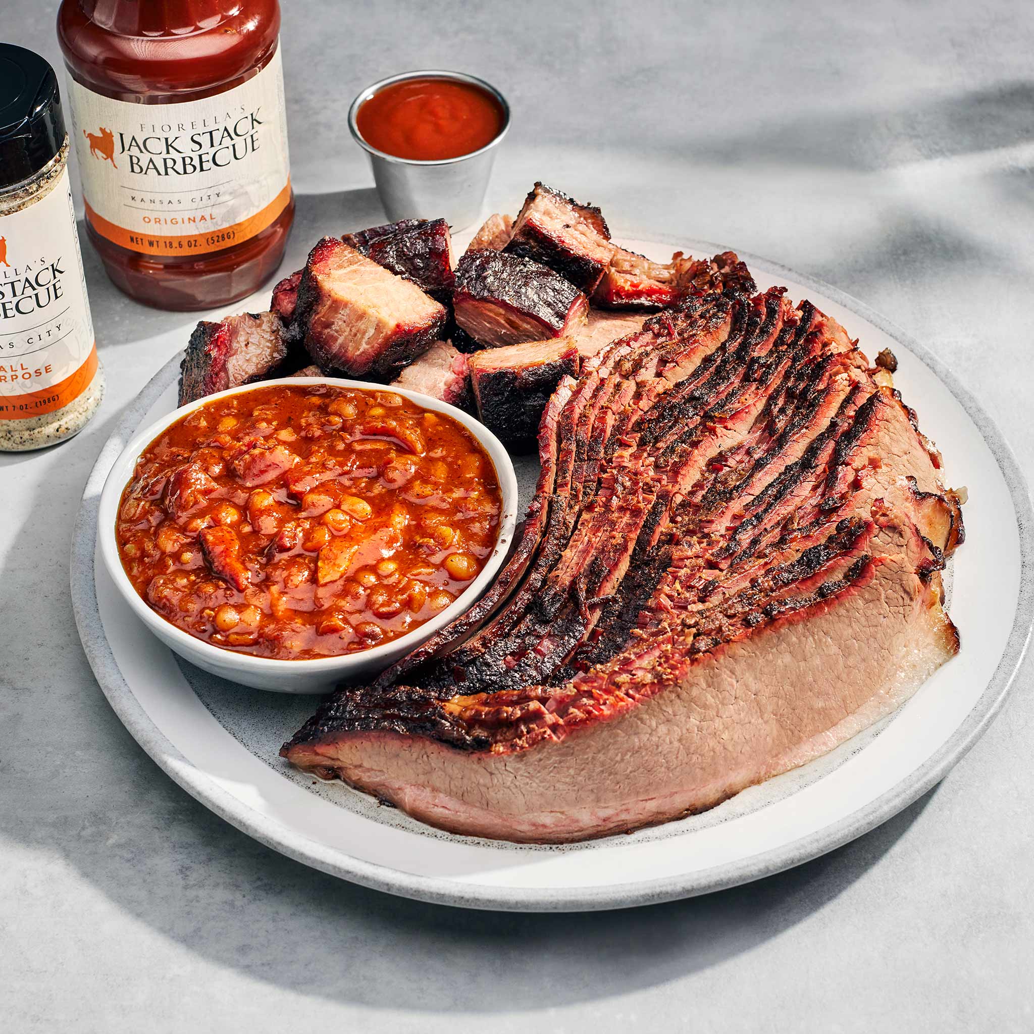 Plated barbecue meal with sliced brisket, baked beans, and Jack Stack Barbecue sauce on a gray surface.