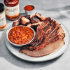 Plated barbecue meal with sliced brisket, baked beans, and Jack Stack Barbecue sauce on a gray surface.