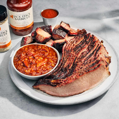 Plated barbecue meal with sliced brisket, baked beans, and Jack Stack Barbecue sauce on a gray surface.