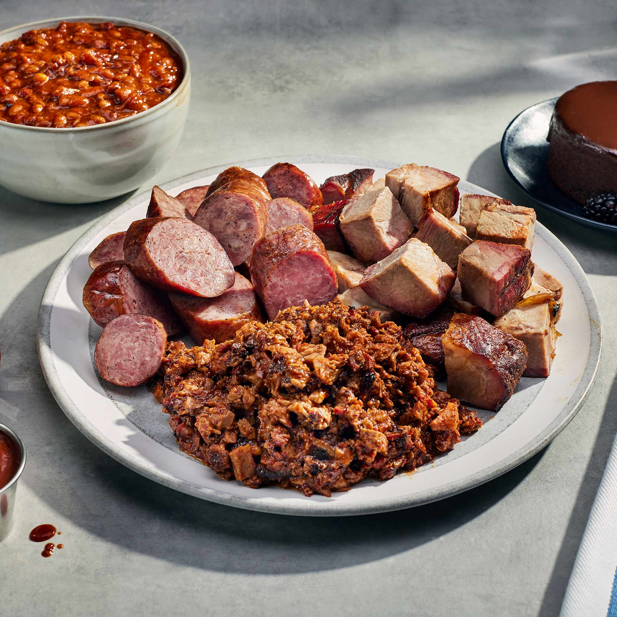Platter of assorted sausages and meatballs on a light surface with a bowl of baked beans in the background.
