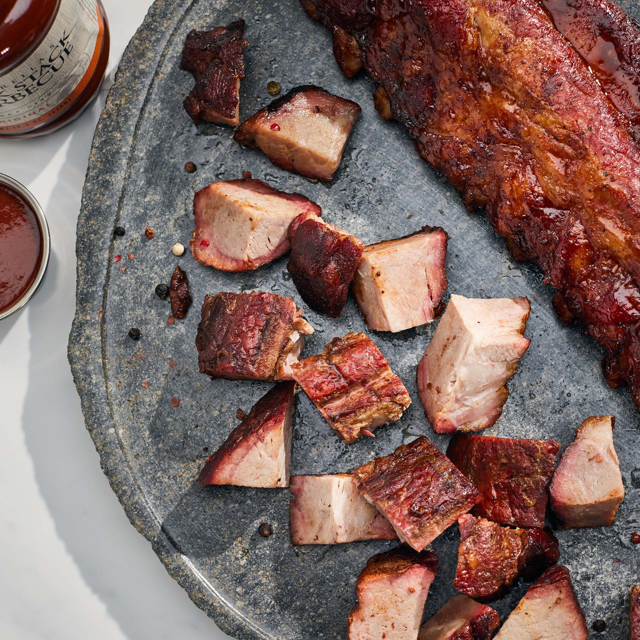 Sliced ribs and meat on a stone platter with a bottle of sauce in the background.