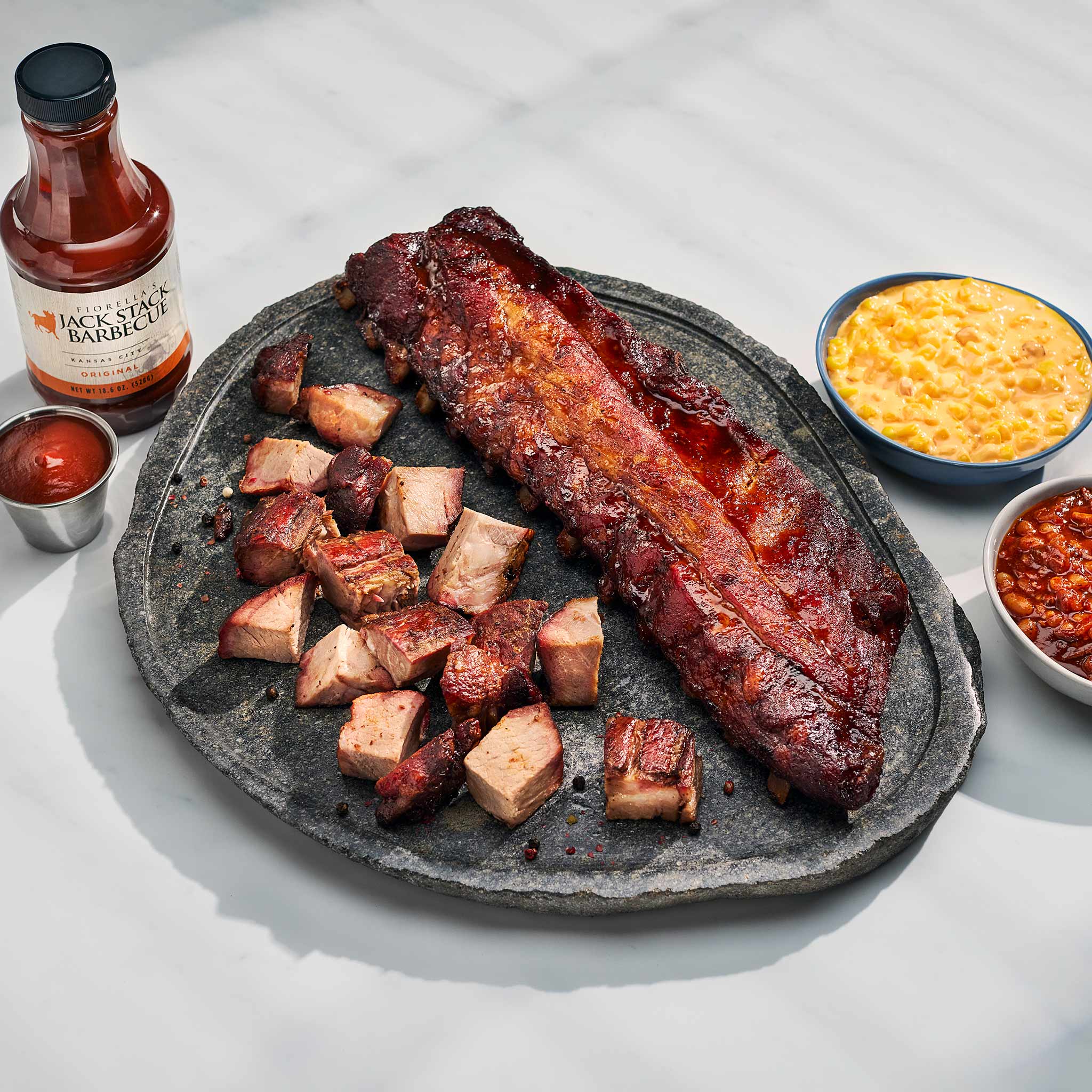 Barbecue ribs and chunks on a stone platter with a bottle of sauce and side dishes.