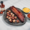 Barbecue ribs and chunks on a stone platter with a bottle of sauce and side dishes.