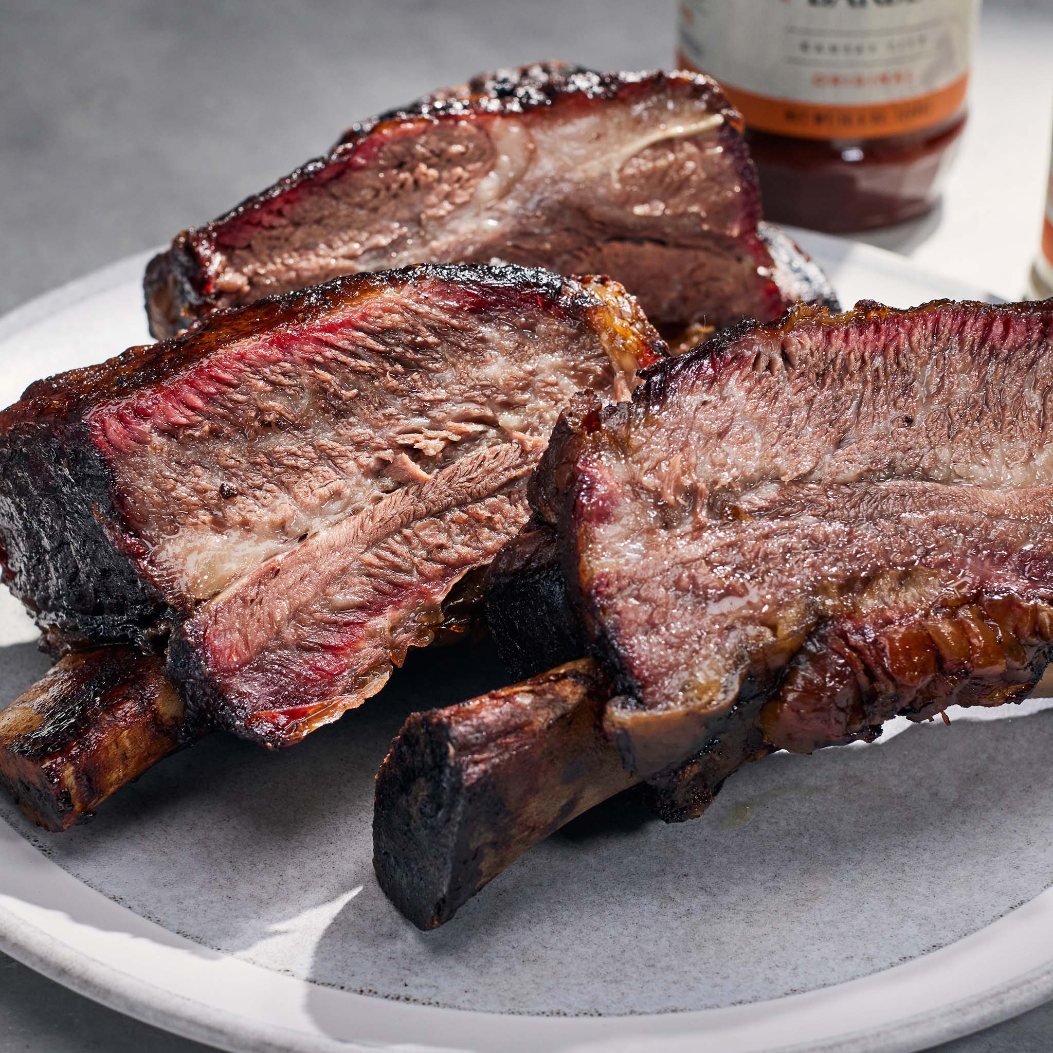 Sliced ribs on a plate with a bottle of barbecue sauce in the background.