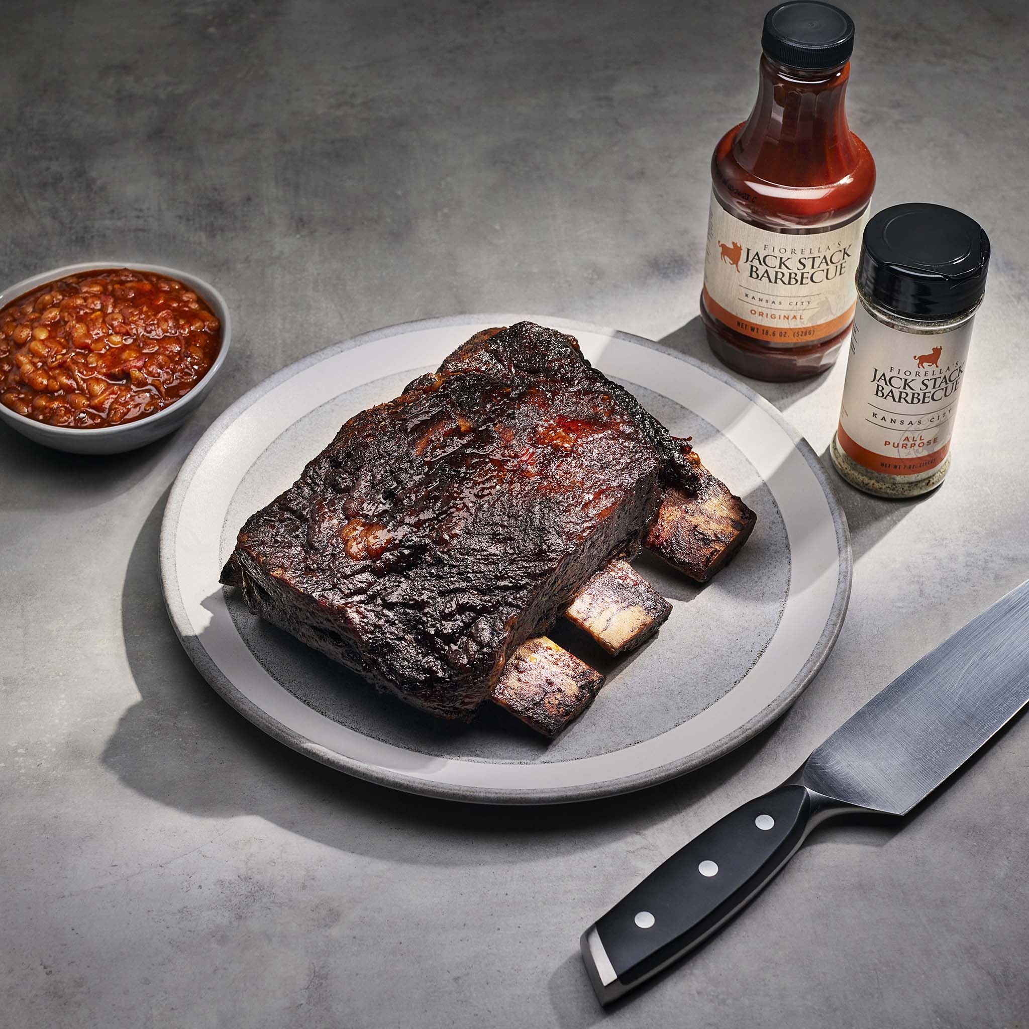 Barbecued ribs on a plate with Jack's Smoke Barbecue sauce and a knife on a gray surface