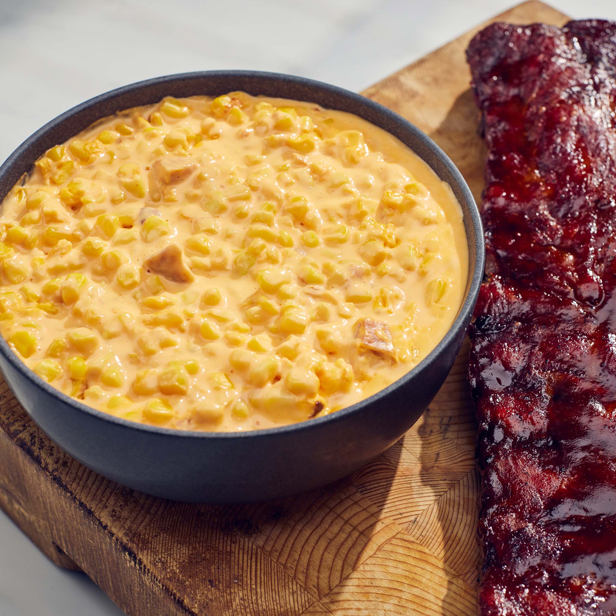 Bowl of corn pudding next to ribs on a wooden cutting board