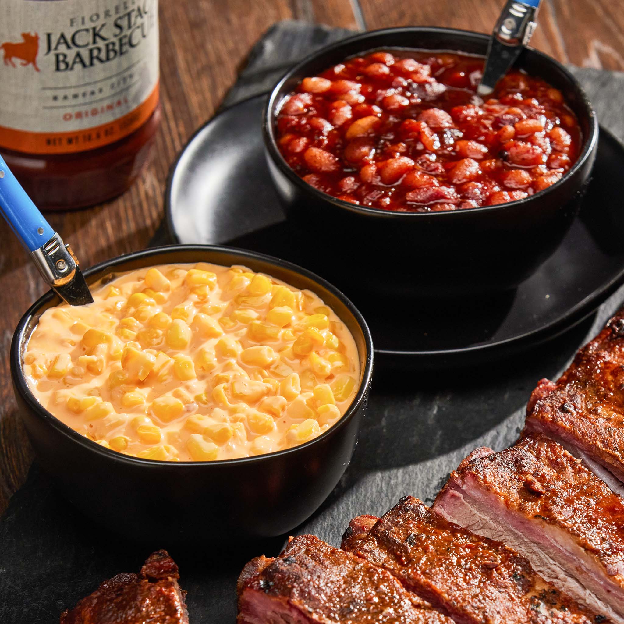 Two bowls of baked beans and corn pudding with smoked meat on a wooden surface, featuring a bottle of Jack Daniel's.
