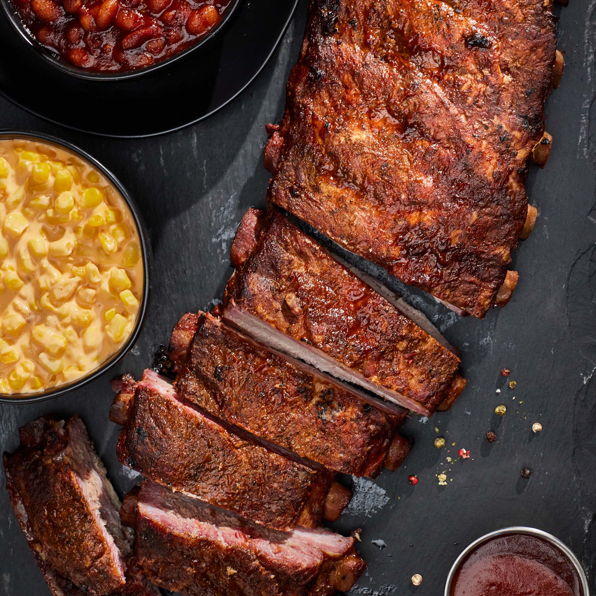 Sliced ribs with sides of baked beans, corn, and macaroni on a dark surface.