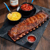 Rack of ribs with sides of baked beans, corn, and ketchup on a slate platter with a bottle of Jack Star's Barbecue sauce.