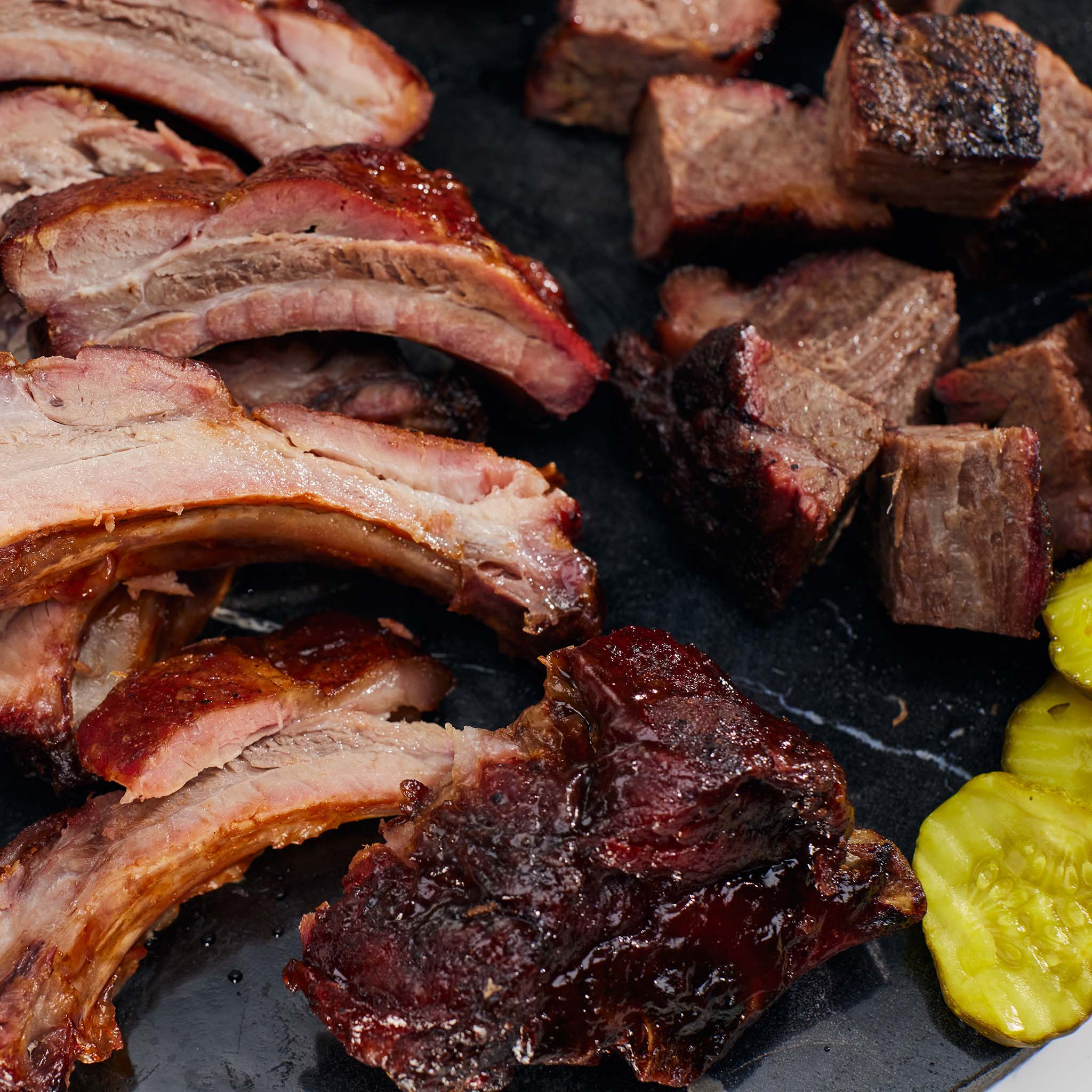 Assorted smoked meats including ribs and chunks on a dark surface with pickles.