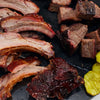 Assorted smoked meats including ribs and chunks on a dark surface with pickles.
