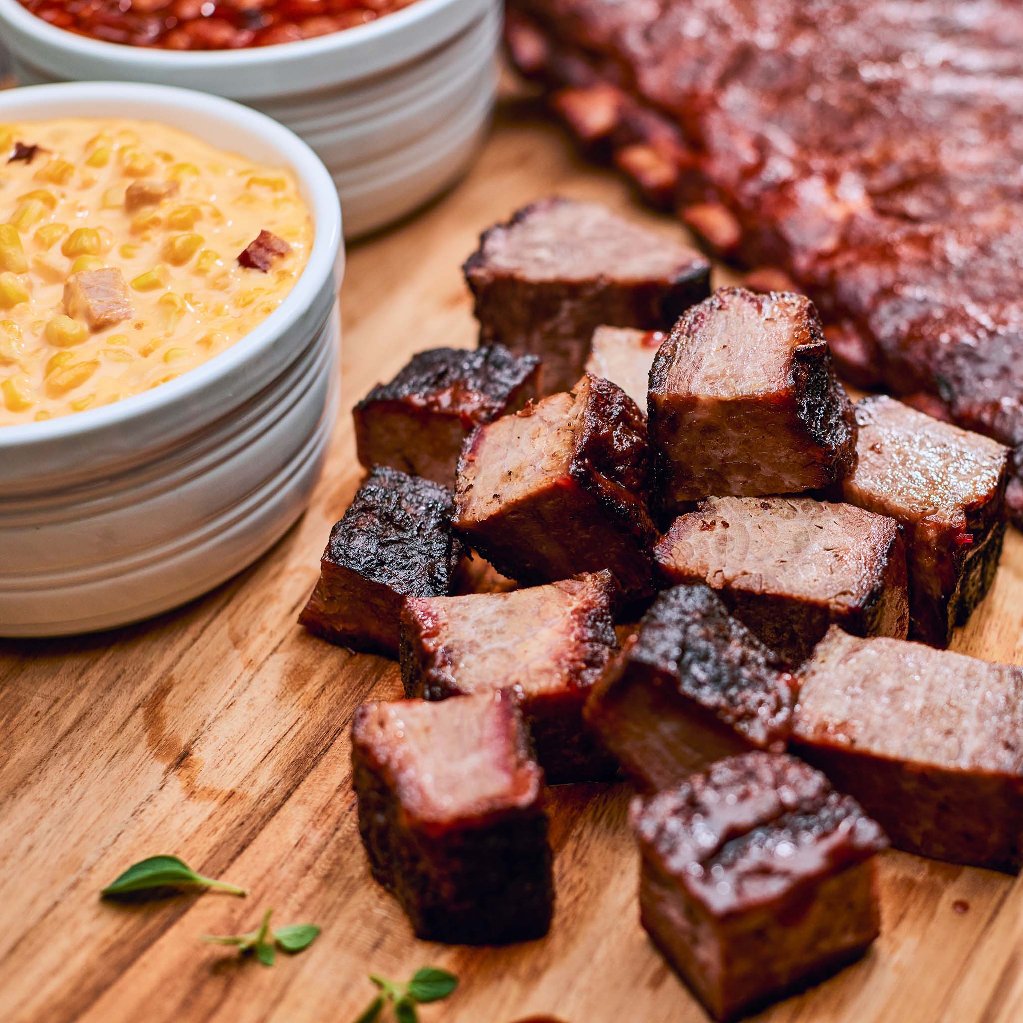 Bbq ribs on a wooden cutting board with bowls of mac and cheese and another dish in the background.