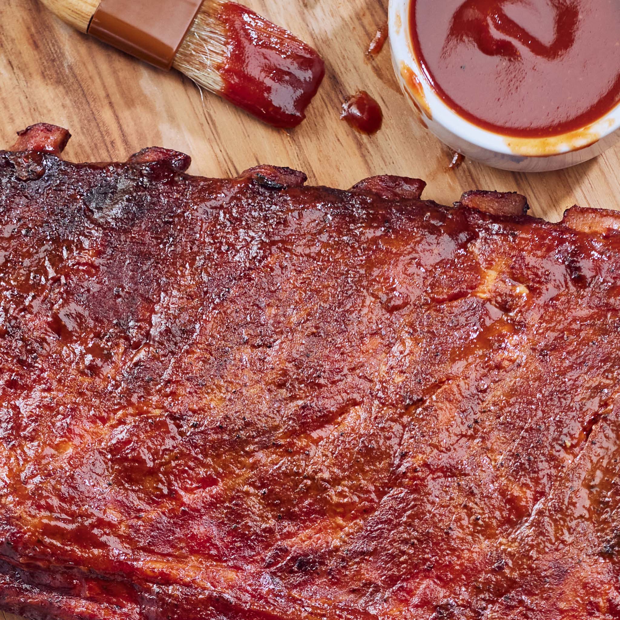 Barbecue ribs with a brush and container of sauce on a wooden surface