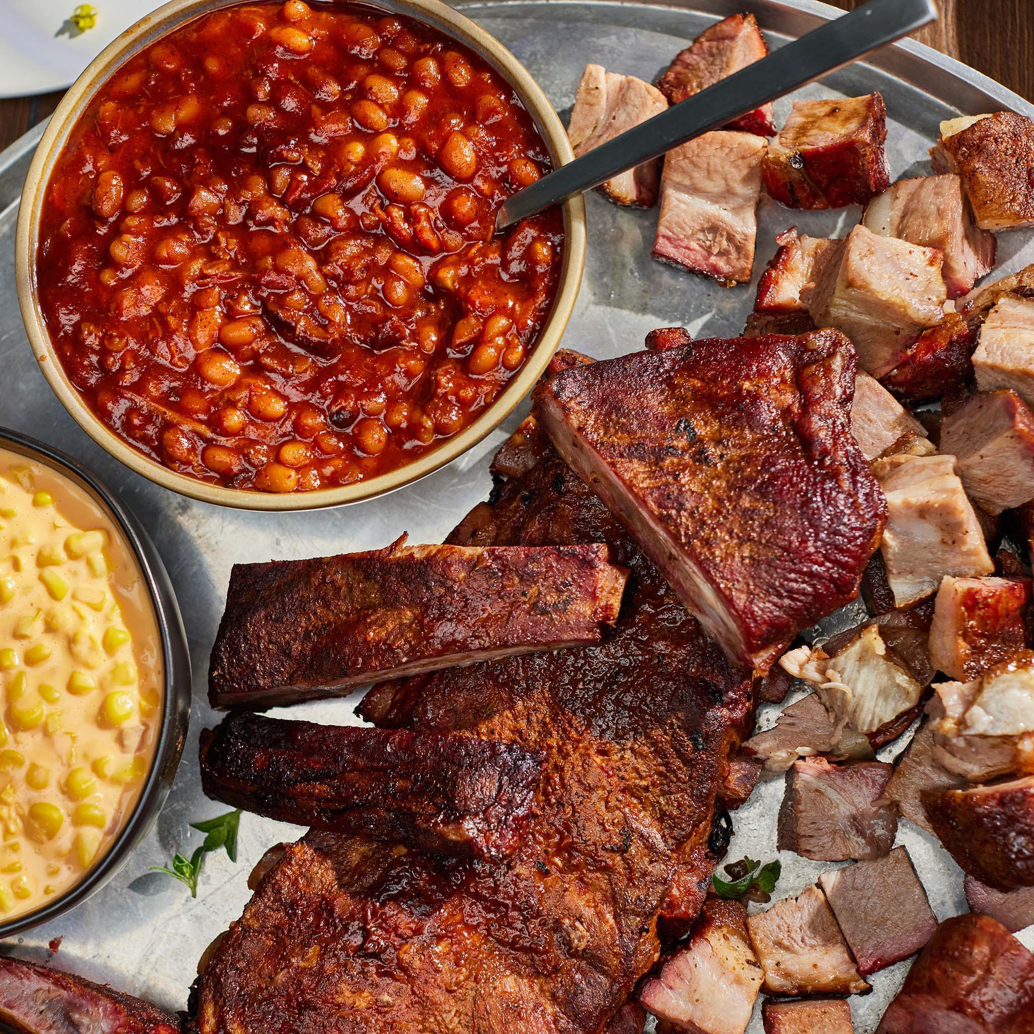 Platter of barbecue meats with a bowl of baked beans and corn on a gray surface.
