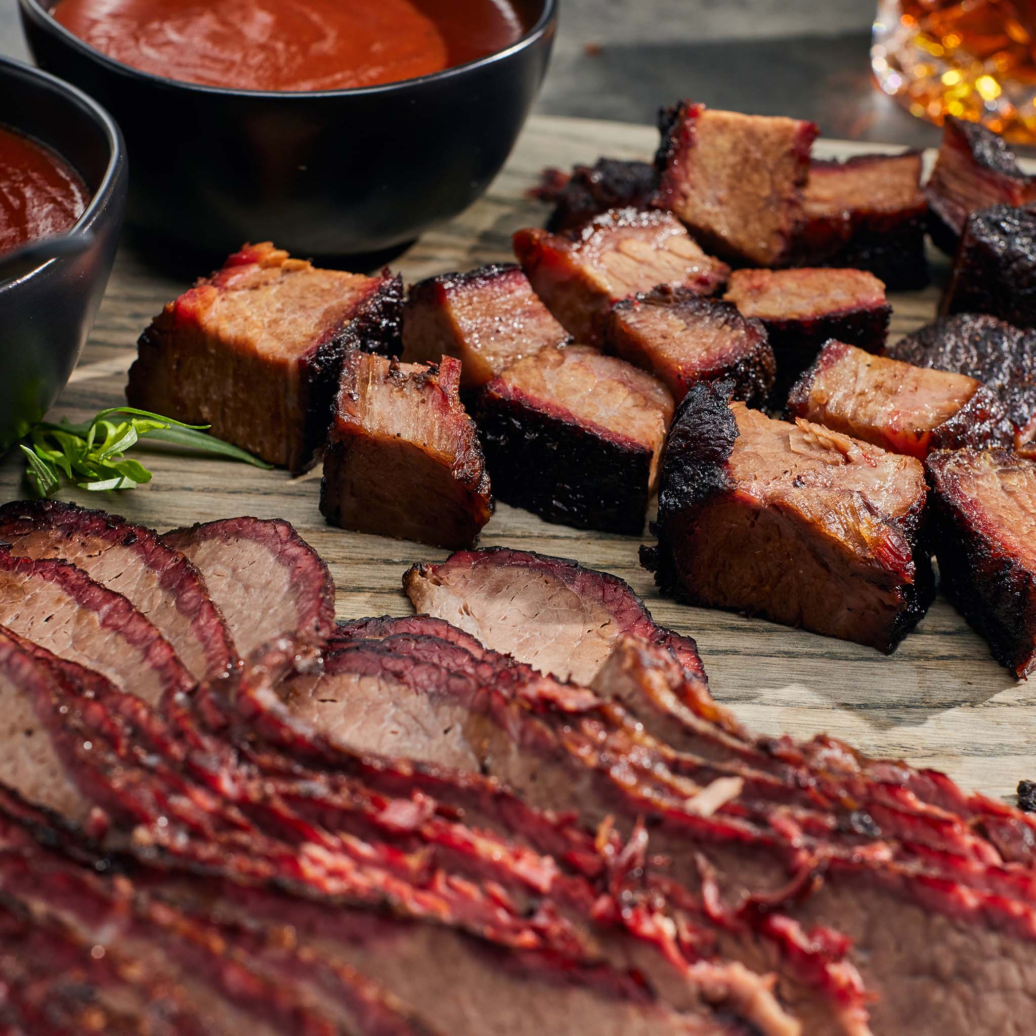 Sliced smoked meat on a wooden board with bowls of sauce in the background