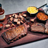 Assorted barbecue meats on a wooden board with sides of mac and cheese and baked beans.