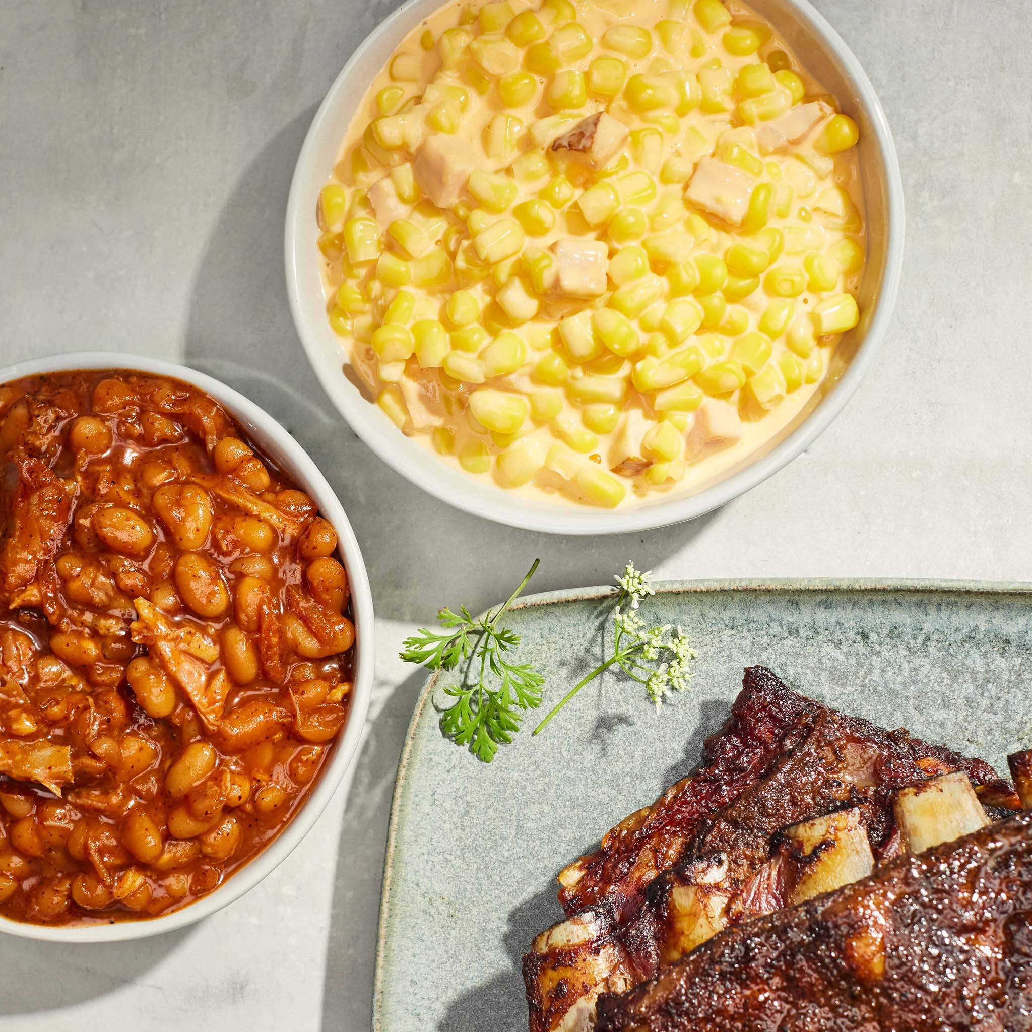 Two bowls of food, one with baked beans and the other with corn, alongside a plate of ribs on a light background.
