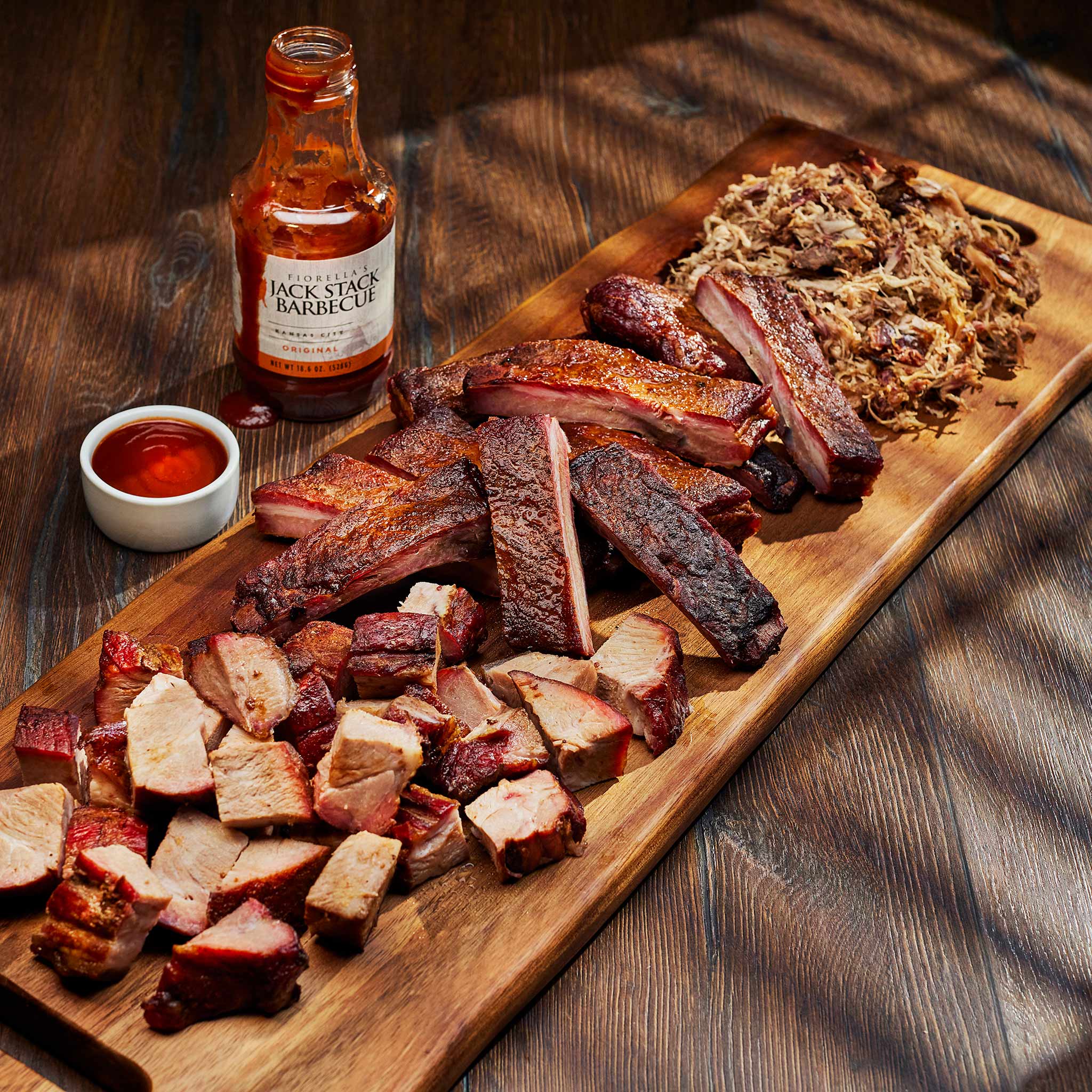 Wooden cutting board with smoked meats and a bottle of Jack Stack Barbecue sauce on a wooden surface.