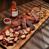 Wooden cutting board with smoked meats and a bottle of Jack Stack Barbecue sauce on a wooden surface.