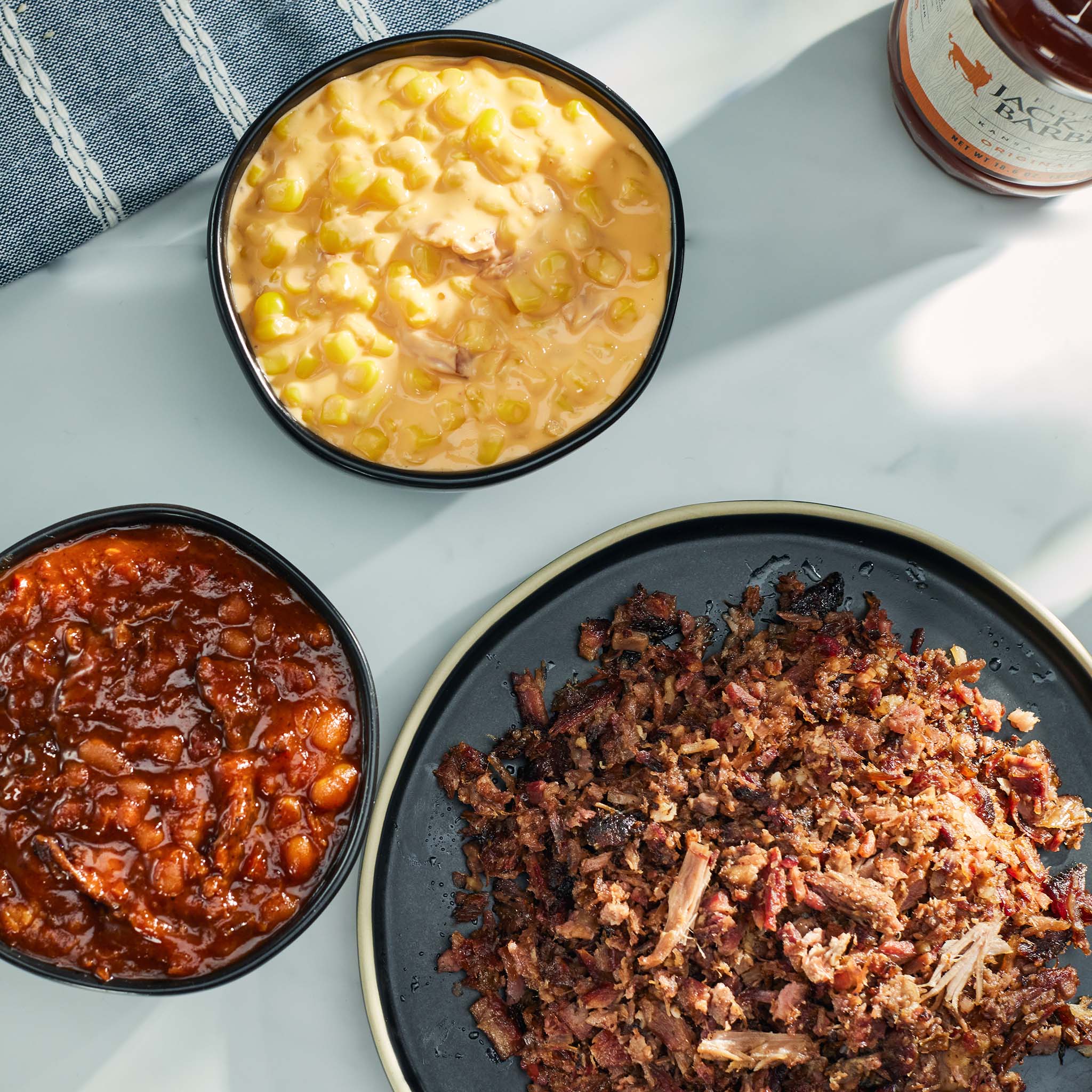 Three bowls of food including corn, baked beans, and pulled pork on a white surface.