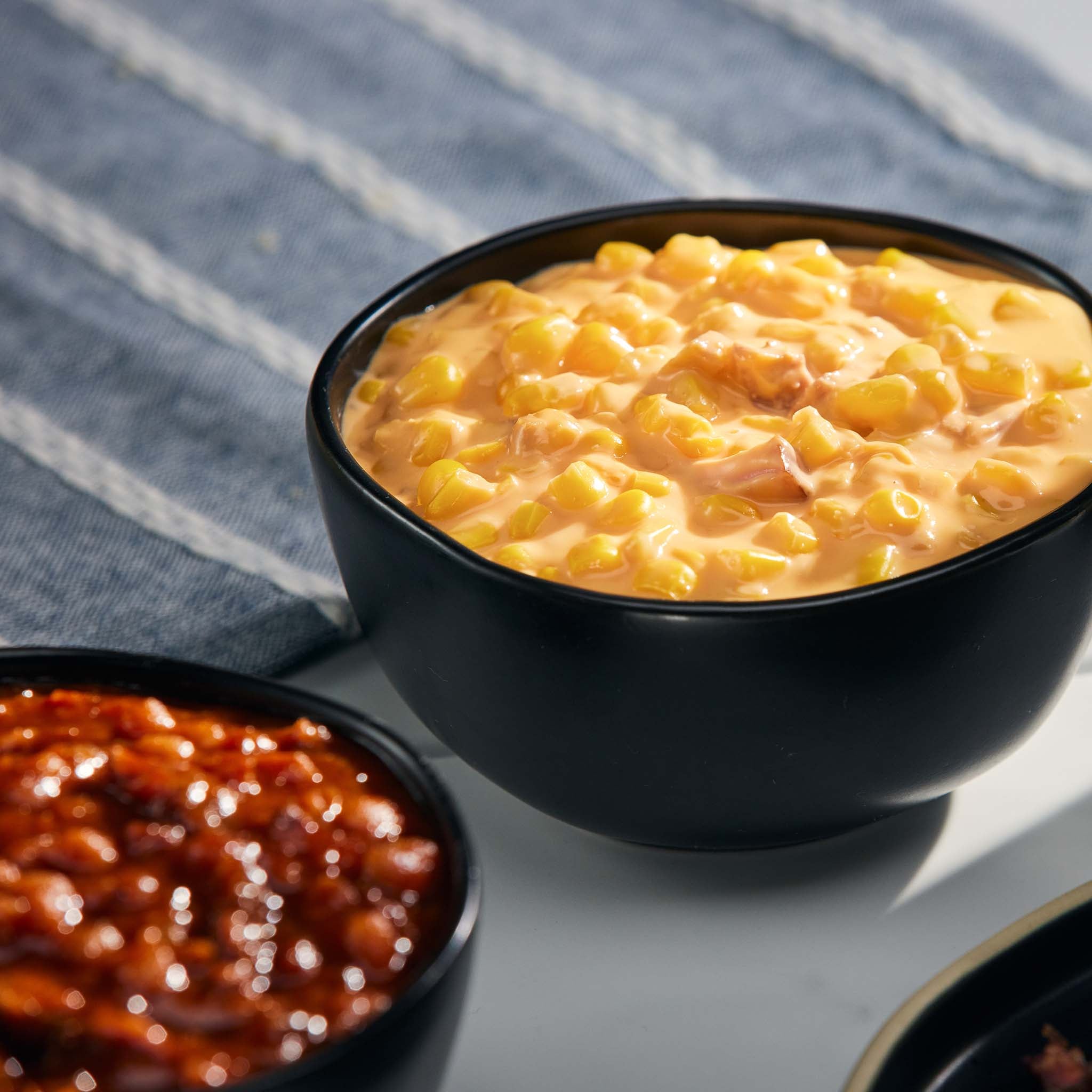 Two black bowls containing cheesy corn and baked beans on a gray surface with a striped fabric background.