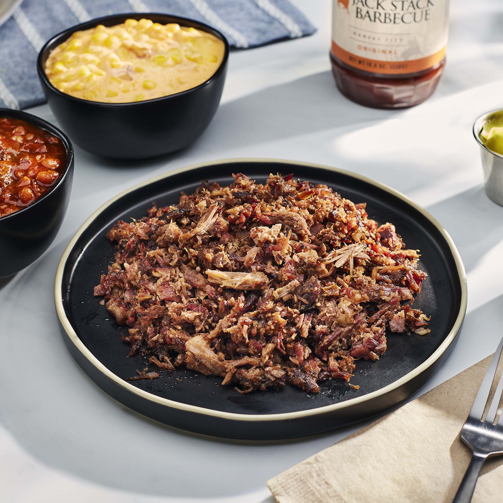 Plated pulled pork with bowls of corn and beans, and a bottle of Jack Stack Barbecue sauce on a light surface.