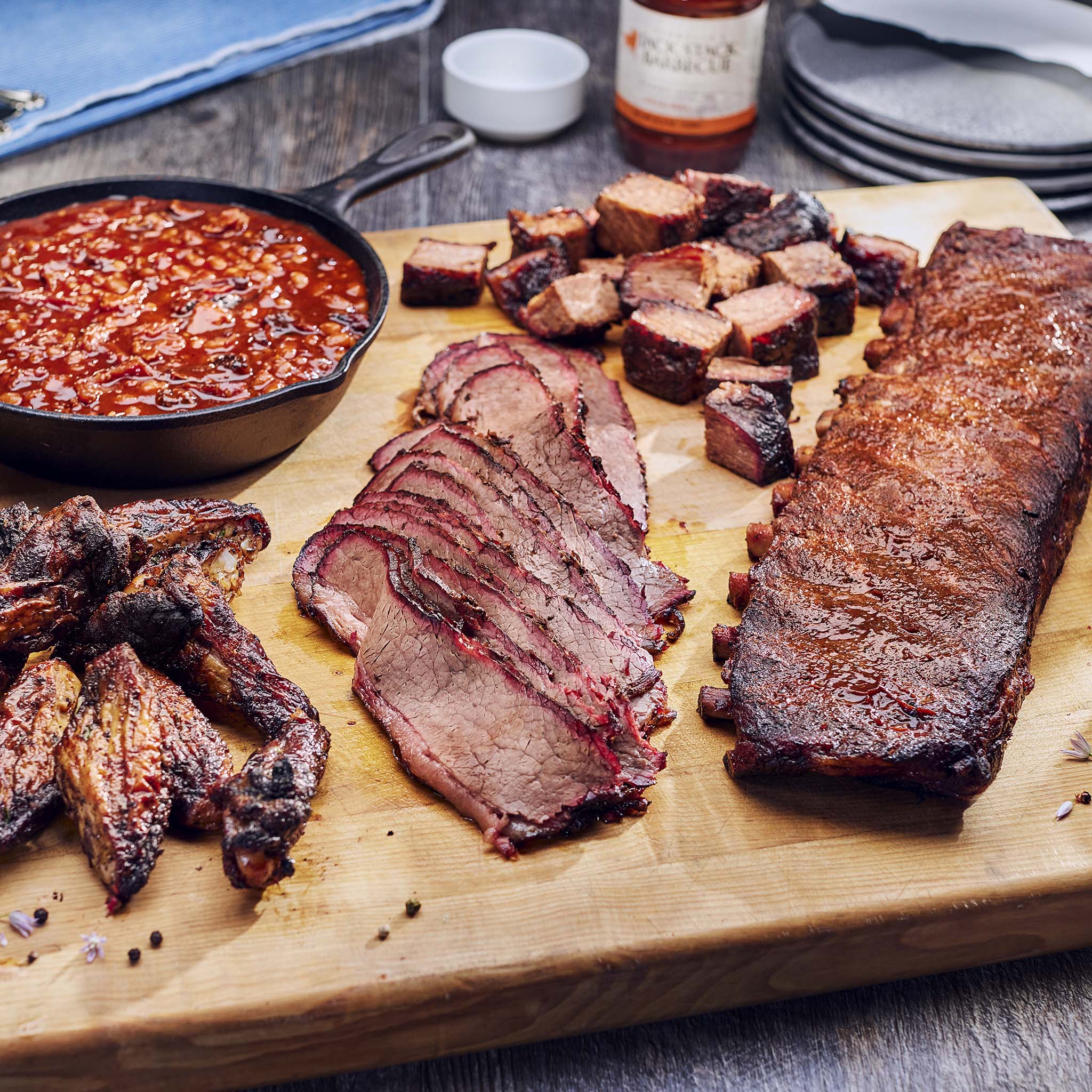 Wooden cutting board with sliced barbecue meats and a bowl of sauce on a rustic wooden table.