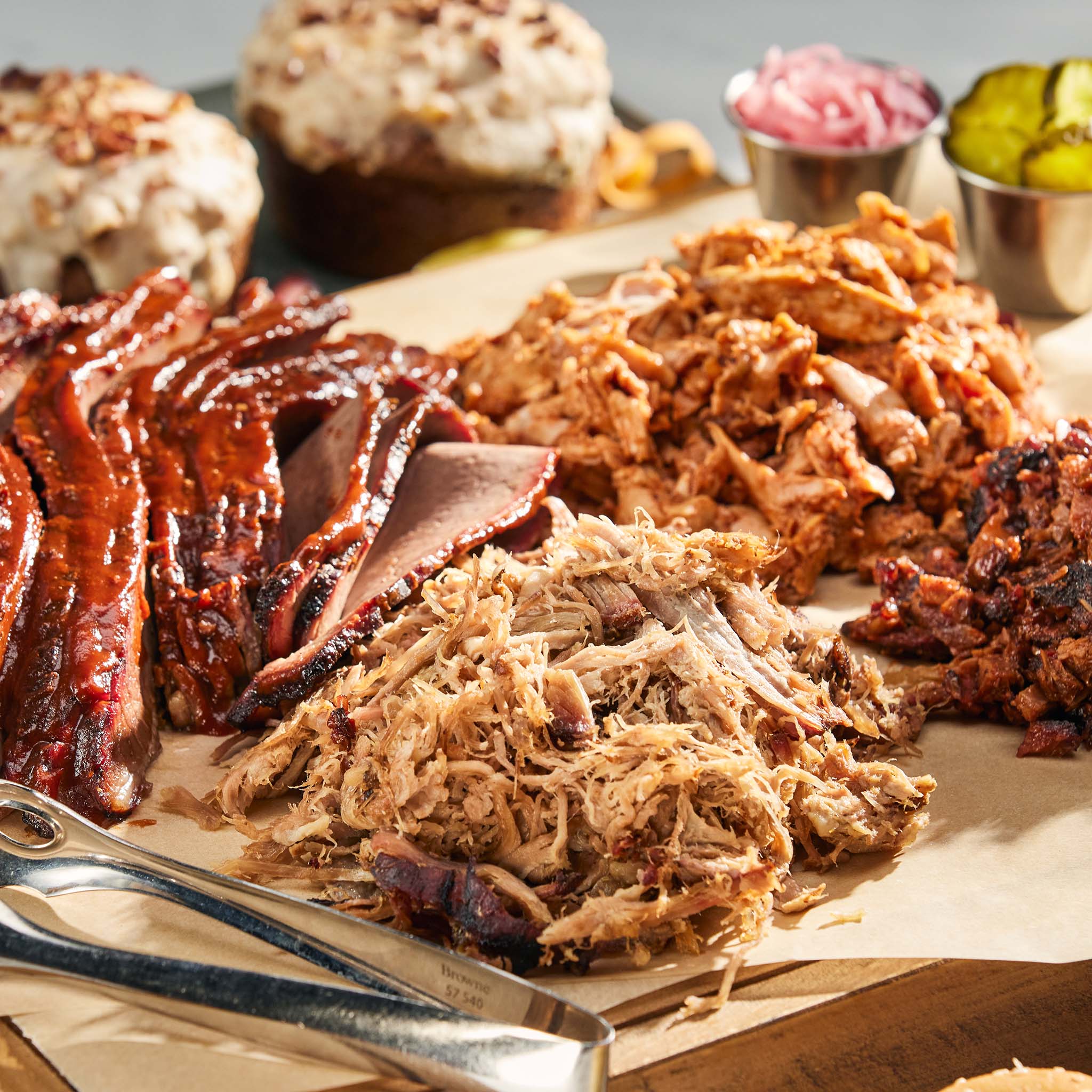 Assorted smoked meats on a wooden cutting board with sides of pickles and coleslaw.