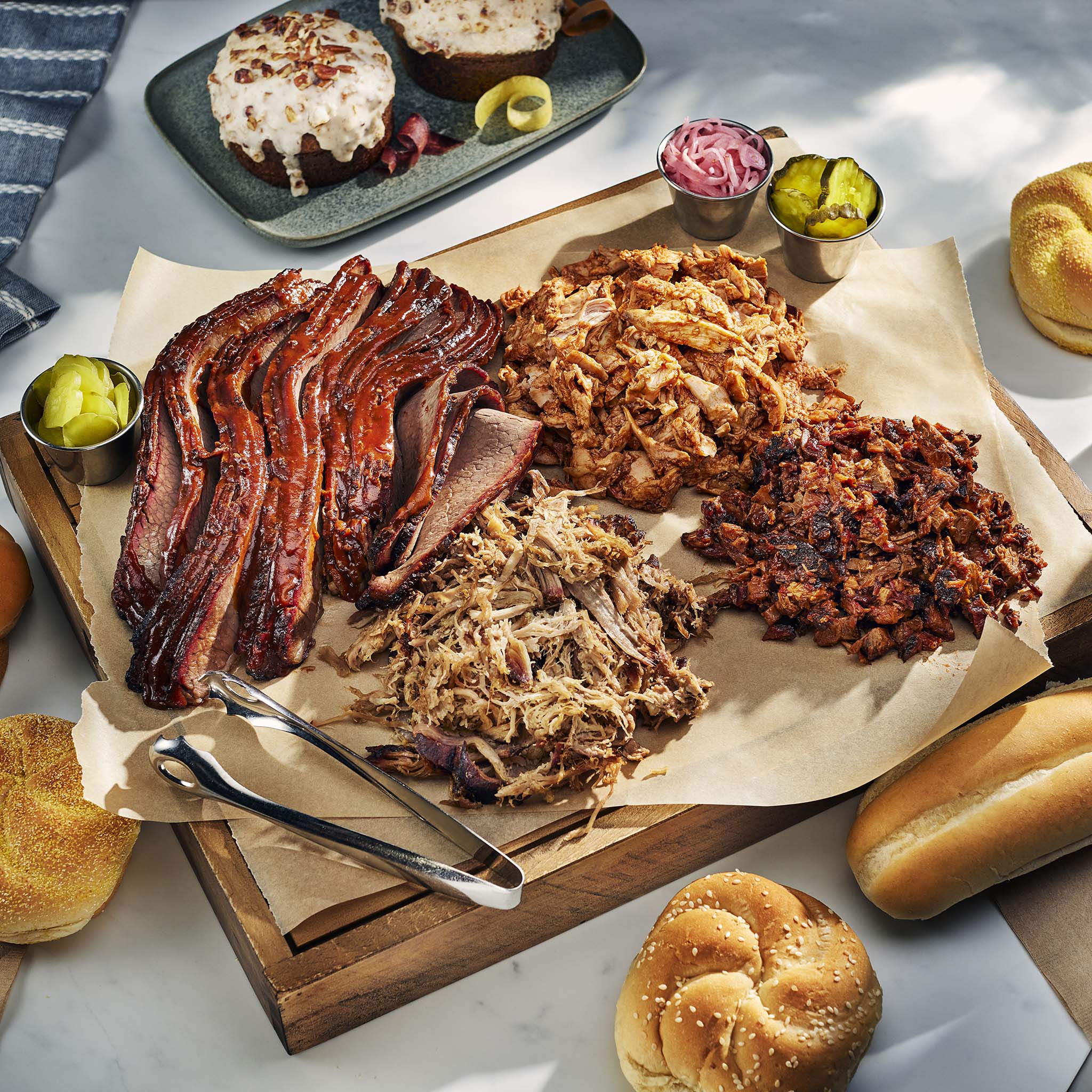 Assorted barbecue meats on a wooden cutting board with sides and buns on a marble surface.