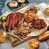 Assorted barbecue meats on a wooden cutting board with sides and buns on a marble surface.