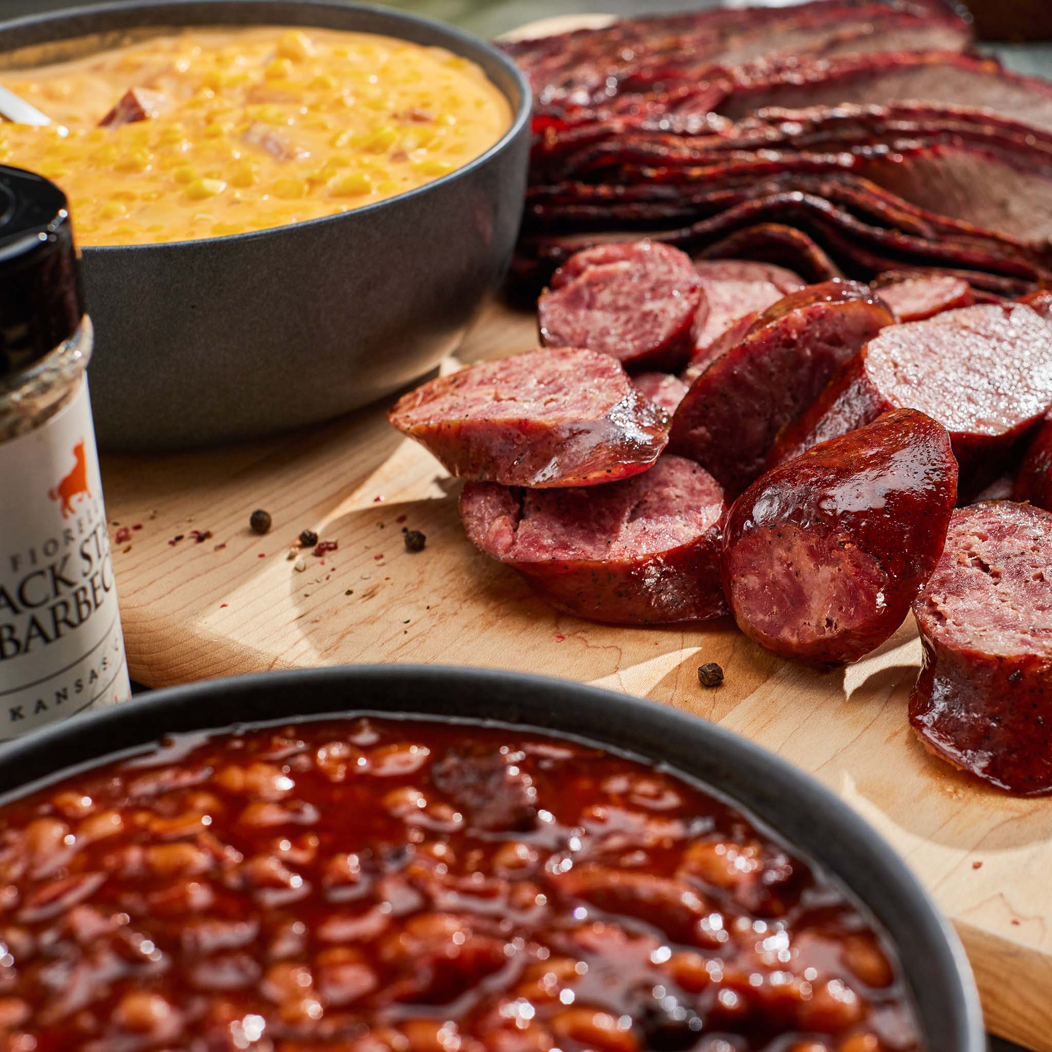 Assorted meats including sliced sausages and bacon on a wooden board with bowls of corn and baked beans.