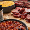 Assorted meats including sliced sausages and bacon on a wooden board with bowls of corn and baked beans.