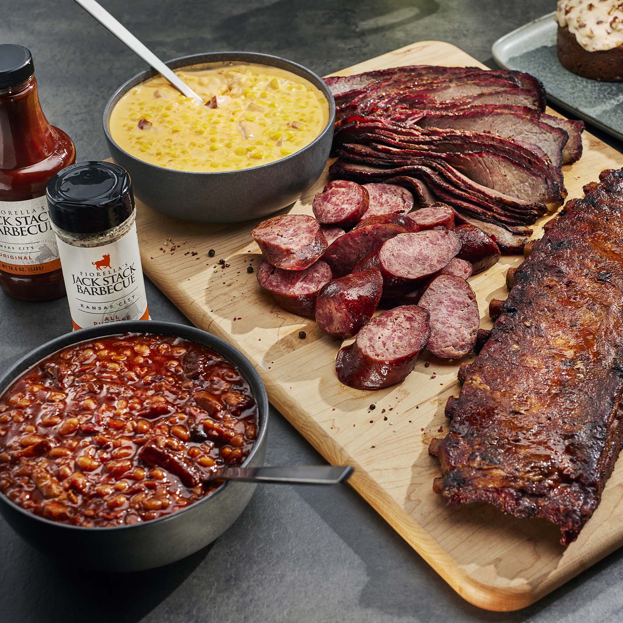 Assorted barbecue meats including ribs, sausages, and corn on a wooden board with barbecue sauce and cornbread.