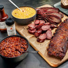 Assorted barbecue meats including ribs, sausages, and corn on a wooden board with barbecue sauce and cornbread.