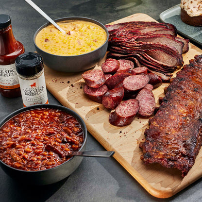 Assorted barbecue meats including ribs, sausages, and corn on a wooden board with barbecue sauce and cornbread.