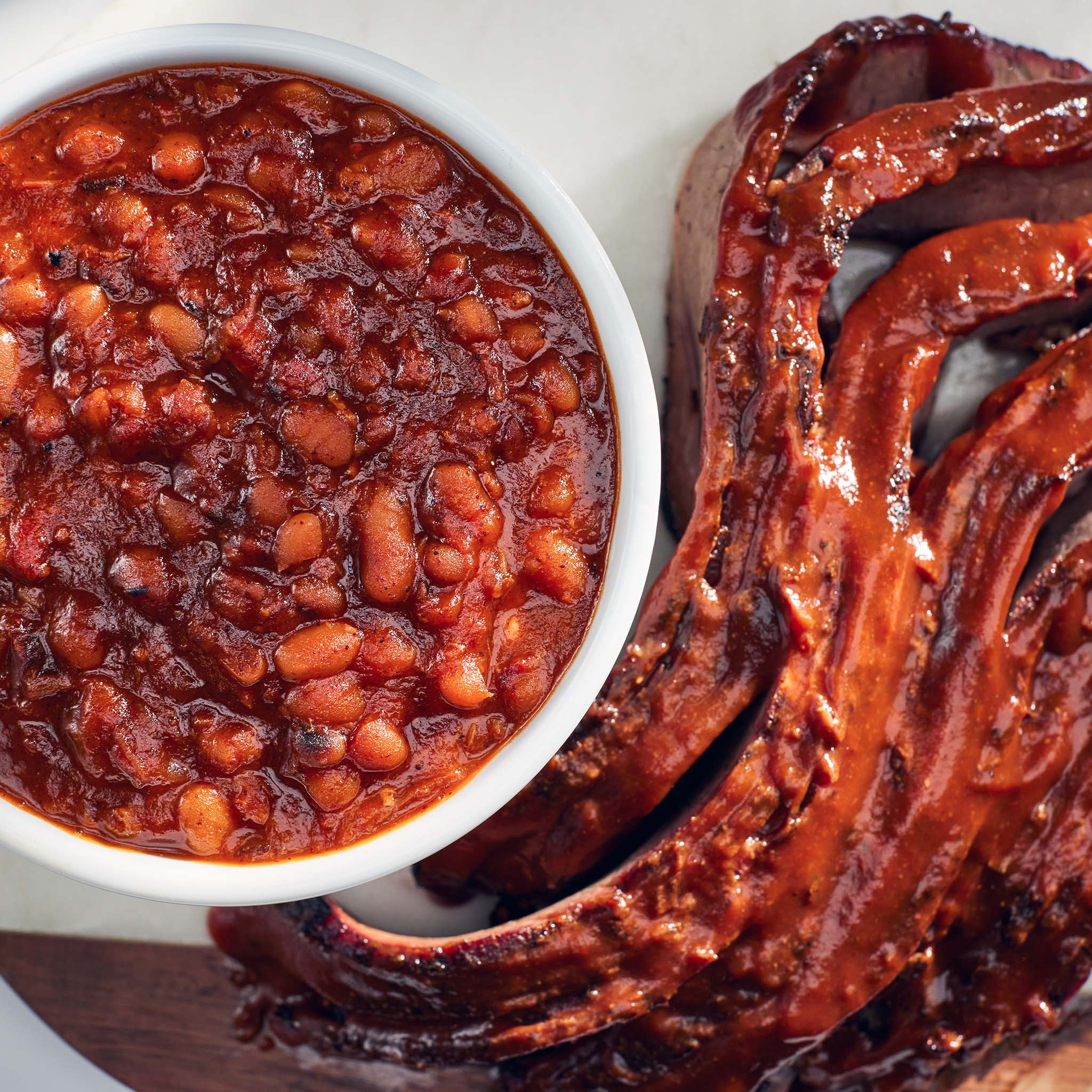 Baked beans in a white bowl with ribs on a white plate