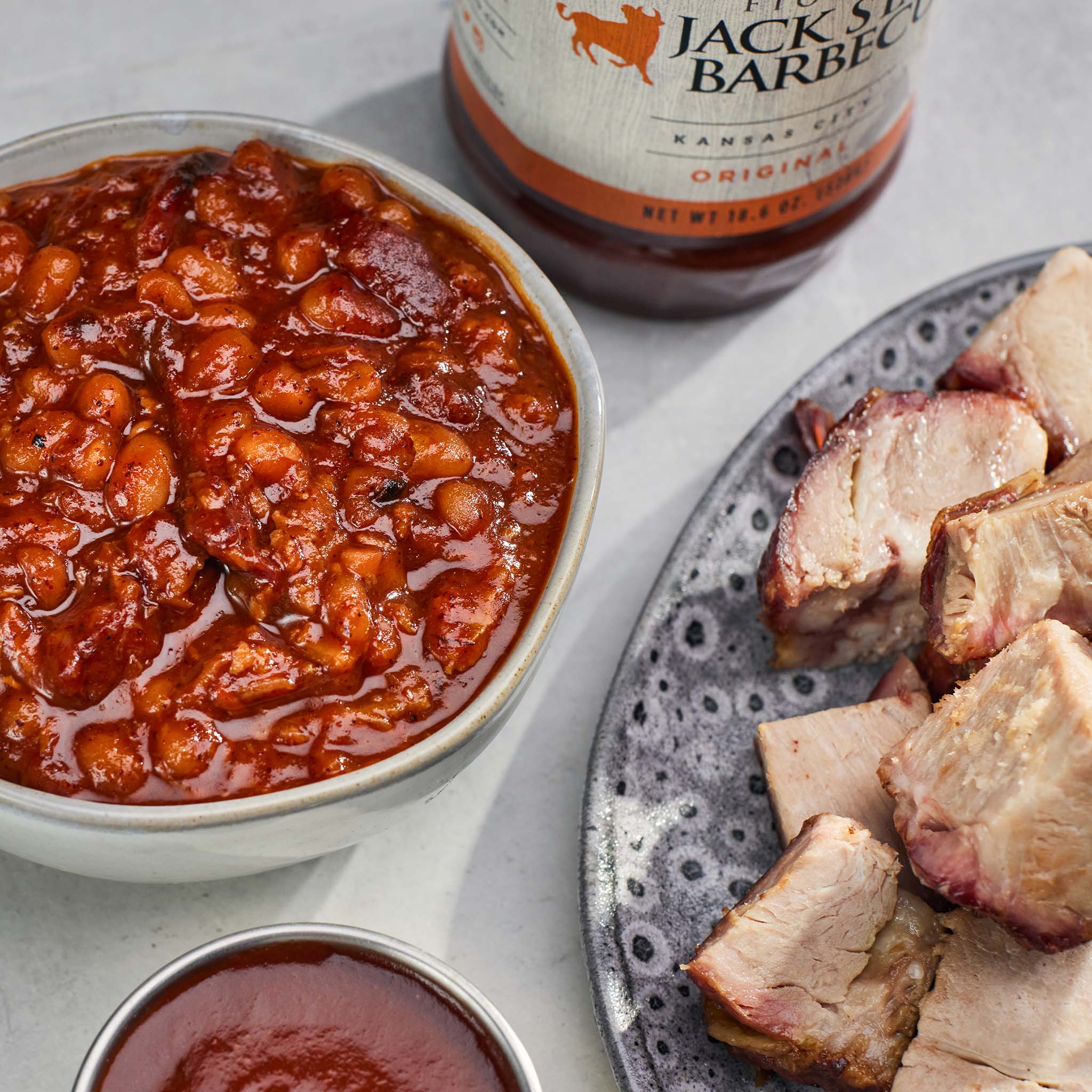 Bowl of baked beans with a jar of Jack Daniel's barbecue sauce and sliced meat on a plate.