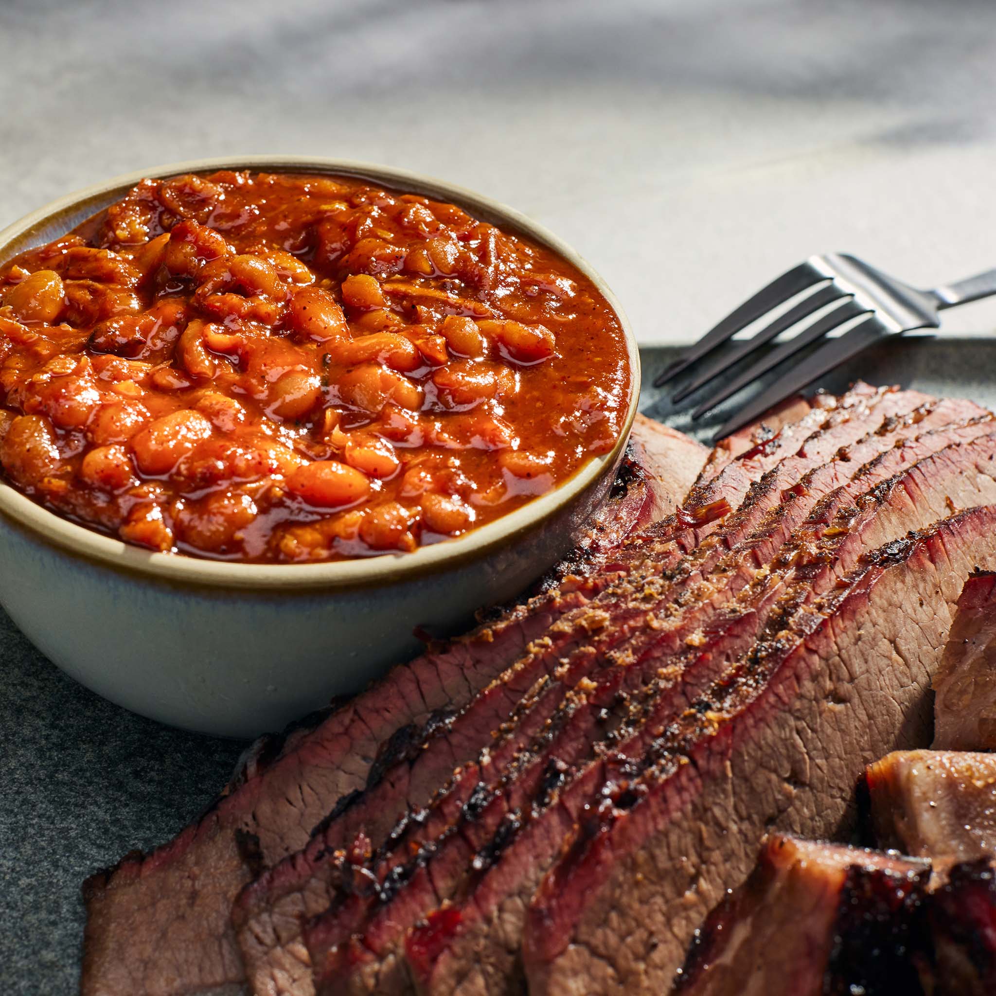 Bowl of baked beans next to sliced brisket on a gray surface