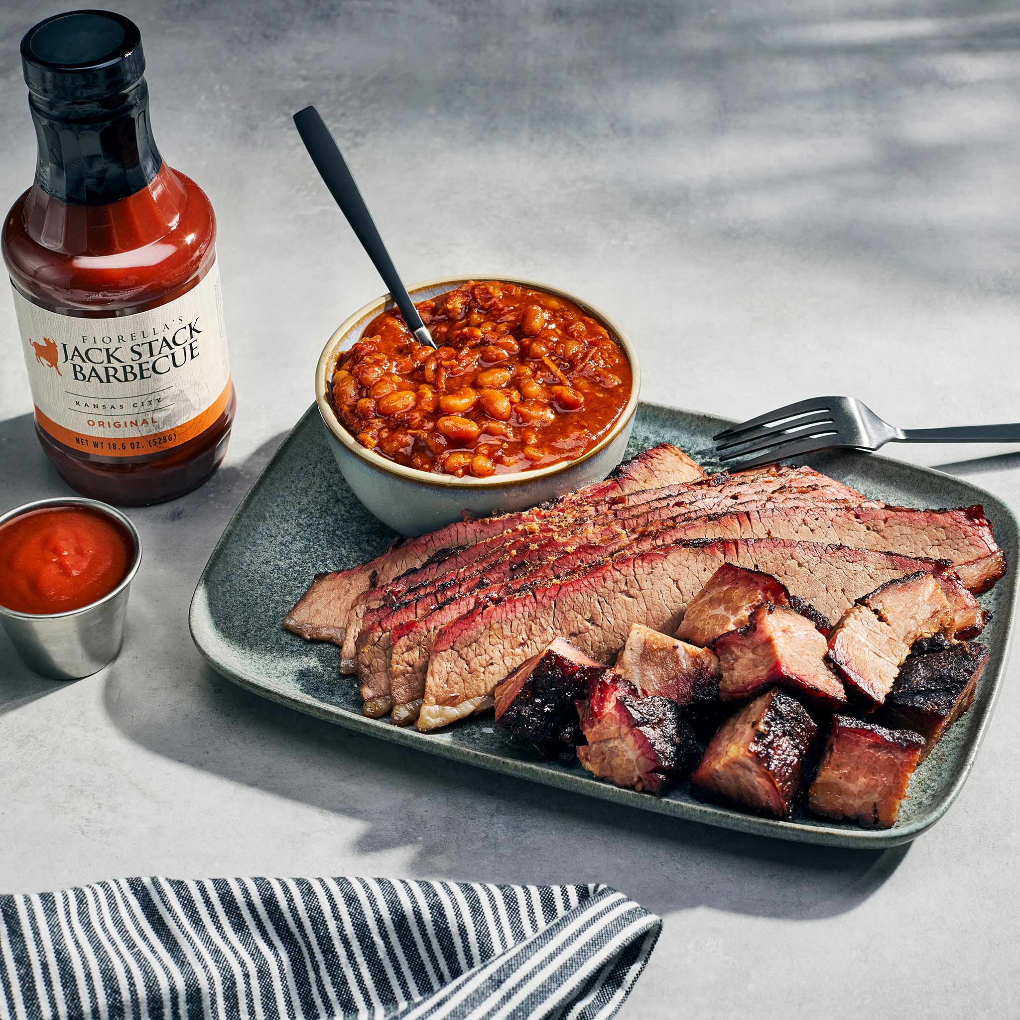 Plated barbecue brisket with a bowl of baked beans and a bottle of Jack Stack Barbecue sauce on a gray surface.