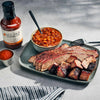 Plated barbecue brisket with a bowl of baked beans and a bottle of Jack Stack Barbecue sauce on a gray surface.