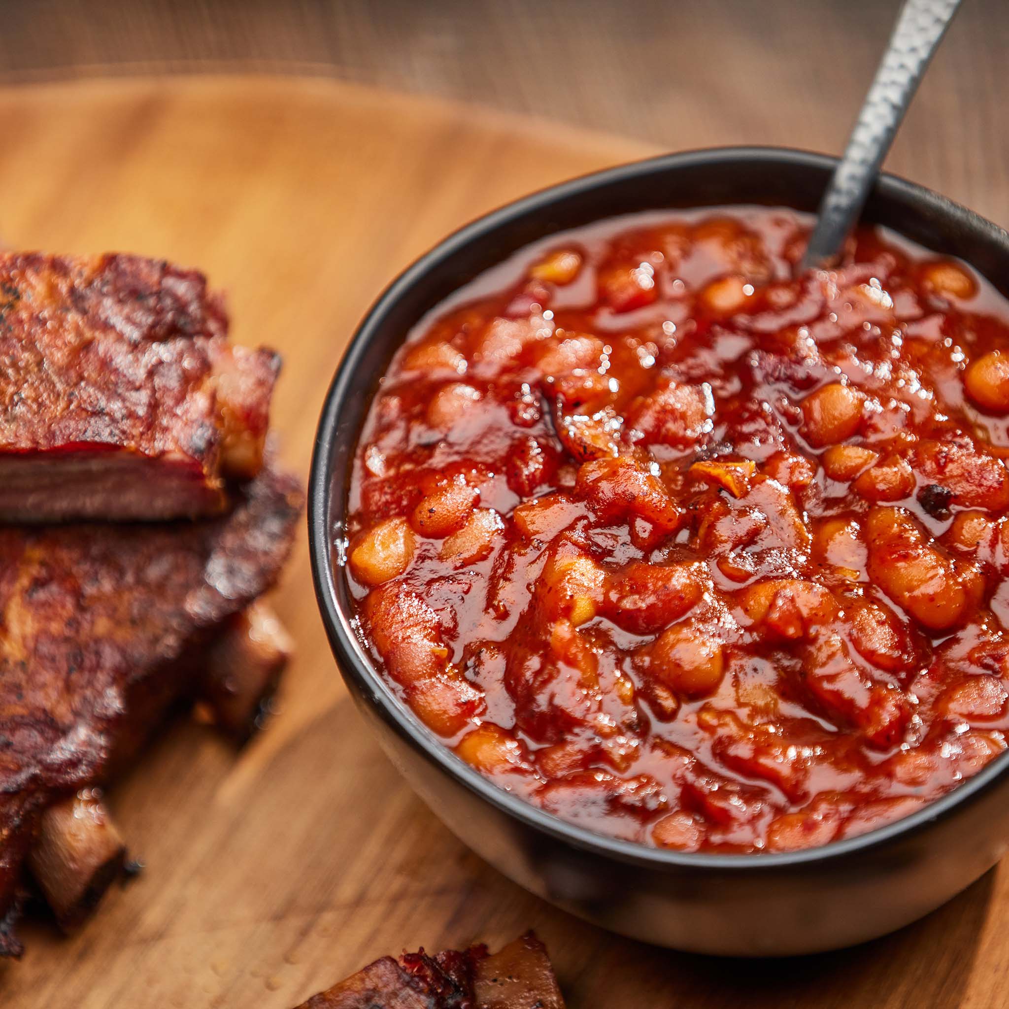 Bowl of baked beans with a spoon on a wooden surface with ribs.
